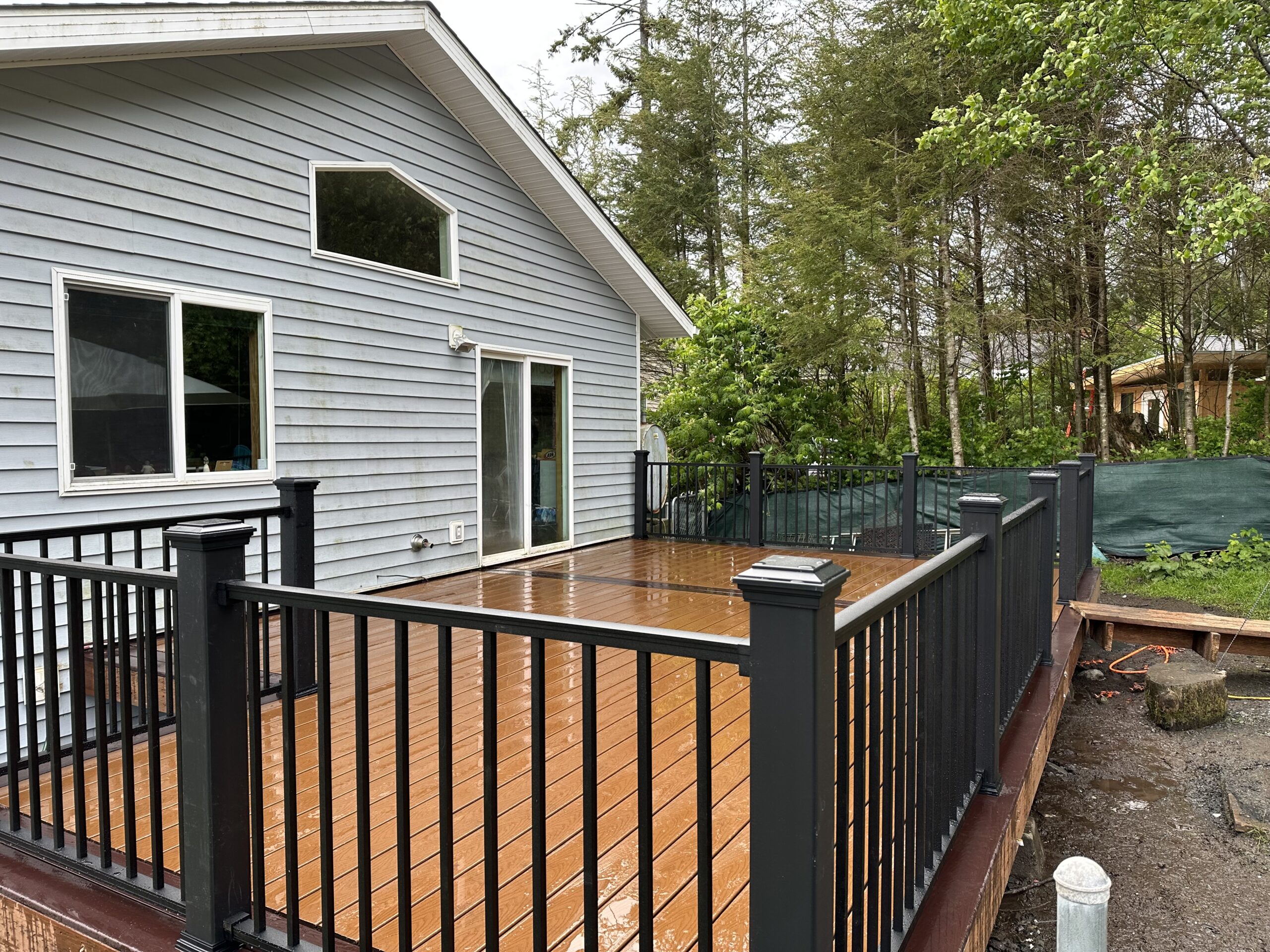 A wet wooden deck with black railings attached to a grey house. There are trees and foliage in the background, and some construction materials on the ground nearby.